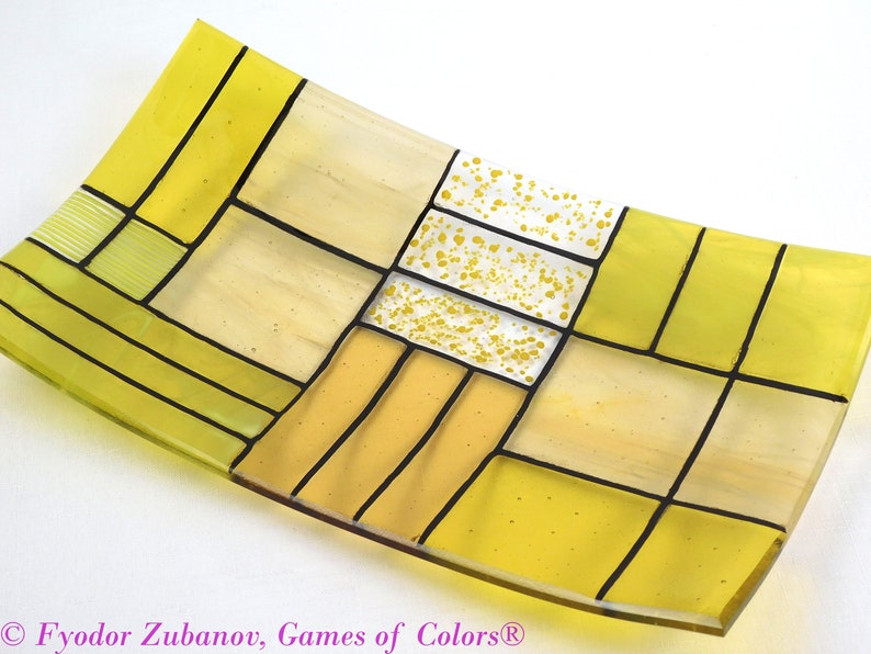 Puede incluir: Un plato rectangular de vidrio con un patr&oacute;n geom&eacute;trico de cuadrados amarillos y crema delineados en negro. El centro del plato tiene un cuadrado blanco con motas amarillas.