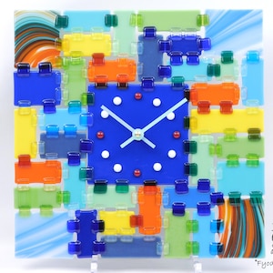 May include: A square, colorful glass clock with a blue face and white hands. The clock face is surrounded by a mosaic of blue, yellow, orange, green, and clear glass pieces. The clock is supported by a clear stand. The text "Games of Colors" and "Fyodor Zubanov" are visible.