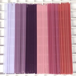 Könnte beinhalten: Eine Sammlung dünner, gerader Bastelstäbchen, die vertikal angeordnet sind. Die Farbpalette reicht von hellem Lavendel über tiefes Lila bis hin zu verschiedenen Rosatönen und endet in einem rötlich-orangen Farbton. Die Stäbchen sind ordentlich ausgerichtet und erzeugen einen ansprechenden Farbverlauf.
