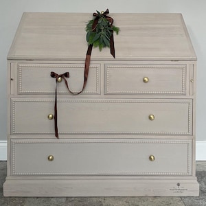 May include: A light grey wooden chest of drawers with four drawers and a top shelf. The dresser has gold knobs and a beaded trim around the drawers. A brown ribbon with a green sprig of greenery is on top of the dresser.