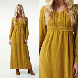 May include: Mustard yellow velvet maxi dress with long sleeves. The dress has a round neckline with three buttons, a fitted waist, and a flowing skirt. The sleeves have a detailed cuff. The model is wearing brown boots.