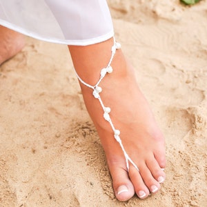 May include: White crochet foot jewelry with white beads on a sandy beach.