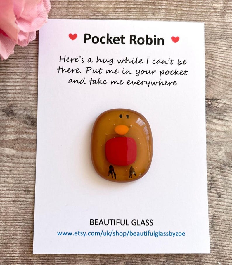 Pu&ograve; includere: Una piccola figurina di pettirosso in vetro con il petto rosso e il corpo marrone. La figurina &egrave; su una carta bianca con il testo "Pocket Robin" e "Here's a hug while I can't be there. Put me in your pocket and take me everywhere".