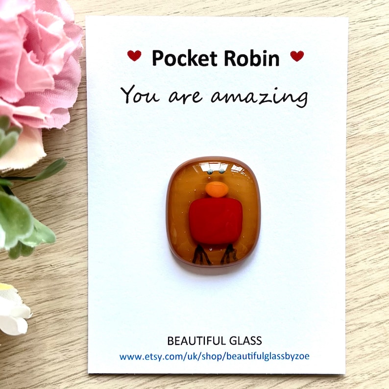 Pu&ograve; includere: Un piccolo pezzo d'arte in vetro a forma di pettirosso con il petto rosso, il corpo marrone e il becco arancione. L'opera d'arte &egrave; su una carta bianca con il testo "Pocket Robin" e "You are amazing".