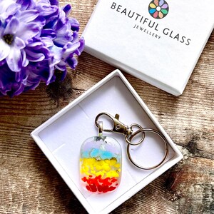 May include: A rectangular glass keychain with a gold-tone clasp and ring. The pendant has layers of red, yellow, blue, and purple glass. The keychain is in a white box with the text "BEAUTIFUL GLASS JEWELLERY".