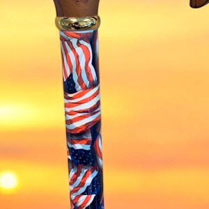 May include: A wooden walking cane with a curved handle and a shaft decorated with American flags. The handle has an engraved American flag. The cane has a gold-colored band near the top.