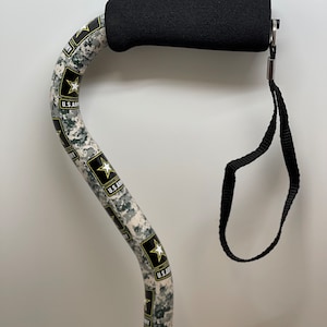 May include: A curved walking cane with a black foam handle and a black wrist strap. The cane's shaft features a camouflage pattern with U.S. Army logos in black and gold.