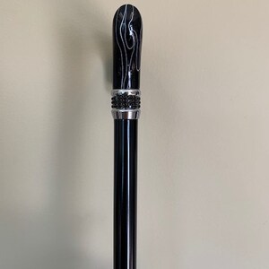 May include: A black walking cane with a decorative handle. The handle is black with white swirling patterns and a silver band with black accents. The shaft is a glossy black.