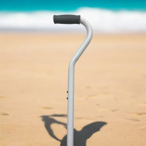 May include: A gray walking cane with a black rubber handle. The cane is standing upright on a sandy beach.