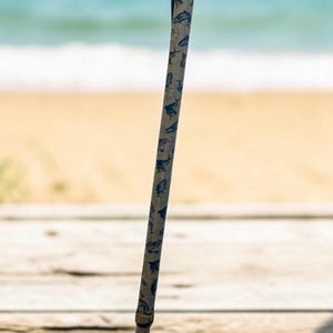 May include: A walking cane featuring a light-colored shaft with a repeating blue fish pattern. The cane has a black ergonomic handle, a gold-colored collar below the grip, a metal adjustment ring in the middle, and a black rubber tip at the base.