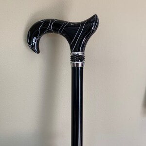 May include: A black walking cane with a curved handle featuring a white swirl design. The handle is connected to a black shaft with a decorative silver band adorned with small, dark embellishments.