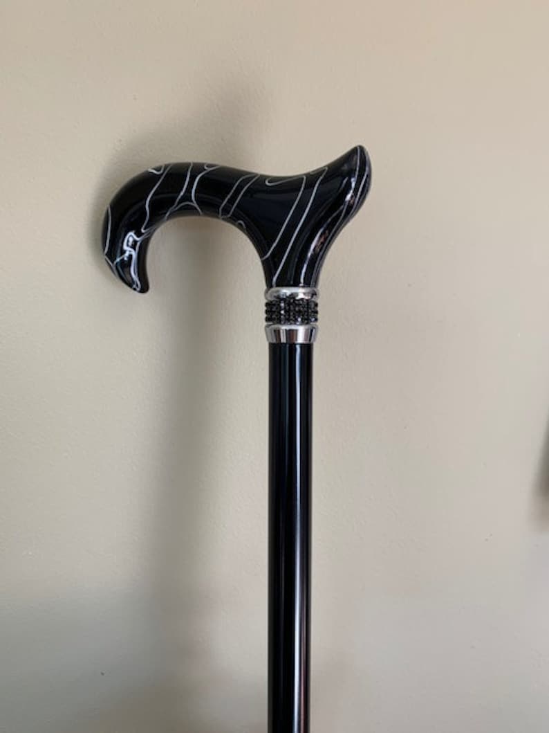 May include: A black walking cane with a curved handle featuring a white swirl design. The handle is connected to a black shaft with a decorative silver band adorned with small, dark embellishments.