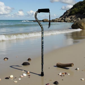 May include: A patterned walking cane with a black handle and wrist strap stands on a sandy beach. The cane has a curved handle and a black rubber tip. The pattern is a mix of blue, green, and white.