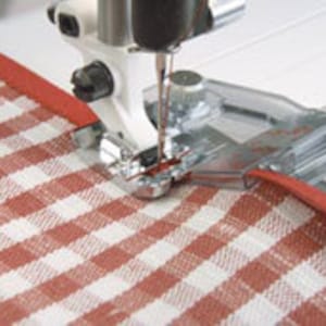 May include: A close-up of a sewing machine foot with a clear plastic attachment for sewing a binding or trim around the edge of fabric. The fabric is a red and white checkered pattern.