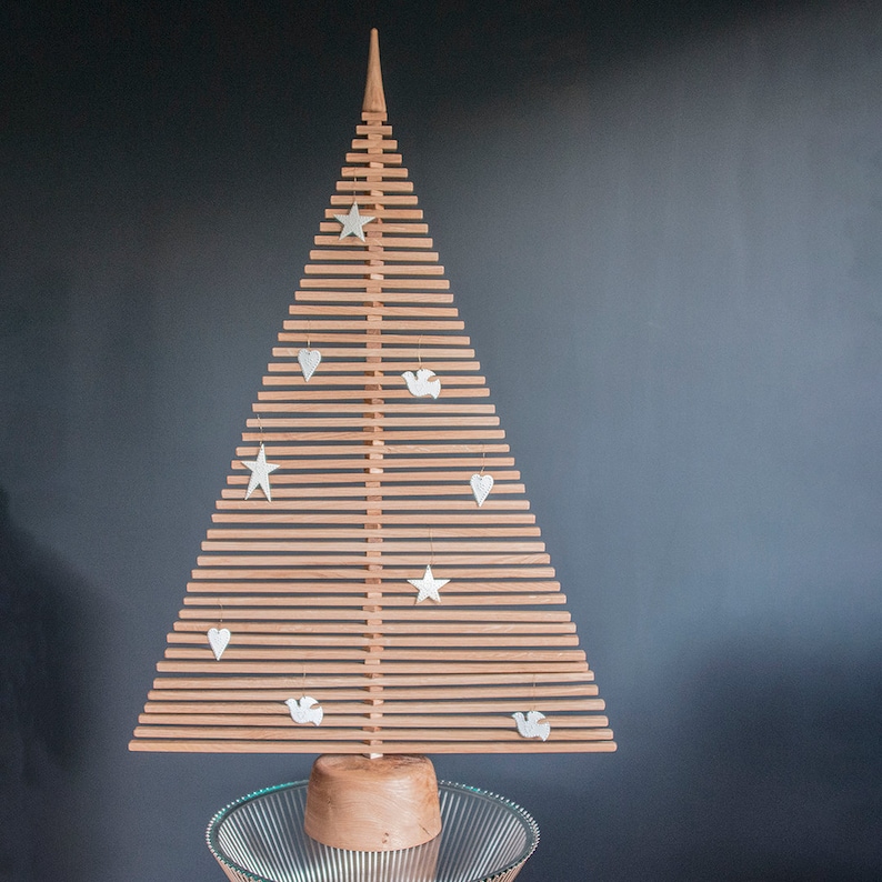 May include: A wooden Christmas tree with a natural wood finish. The tree is decorated with white stars, hearts, and doves. The tree is standing on a clear glass base.