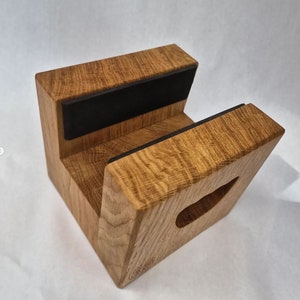 May include: A wooden phone stand with a natural oak finish. The stand has a rectangular base with a cutout and two black rubber pads for device support. The wood grain is visible, and the stand is designed to hold a phone.