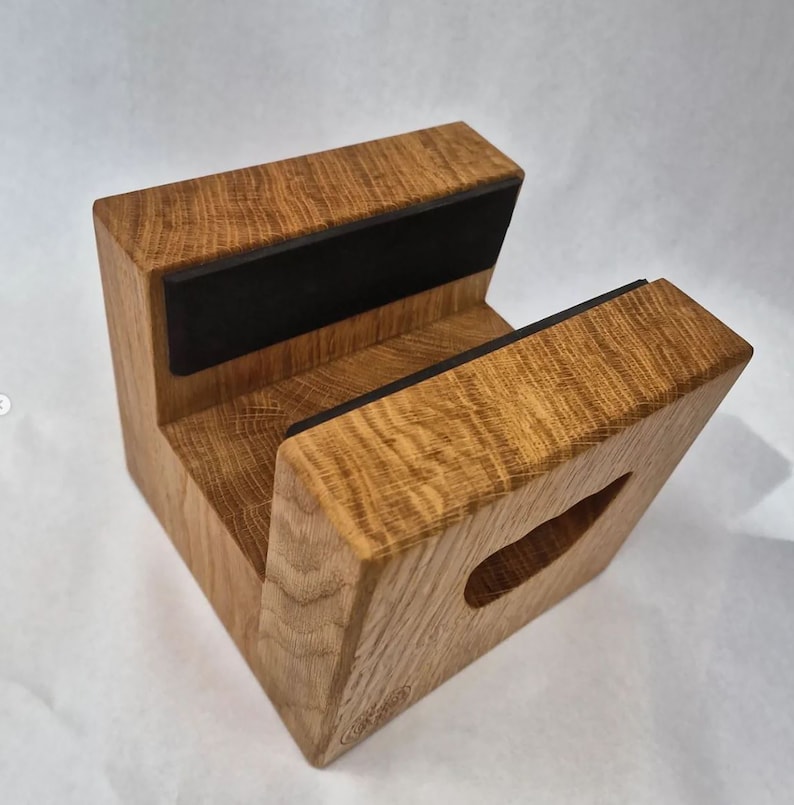 May include: A wooden phone stand with a natural oak finish. The stand has a rectangular base with a cutout and two black rubber pads for device support. The wood grain is visible, and the stand is designed to hold a phone.