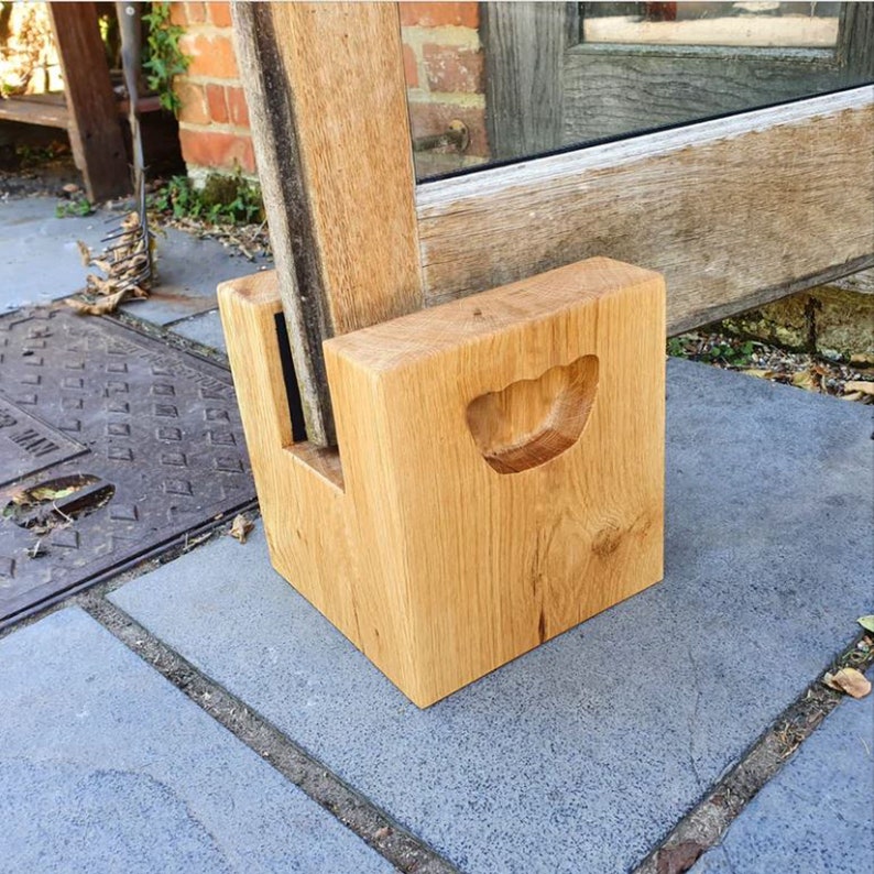 May include: A light-colored wooden doorstop with a carved handle. The doorstop is rectangular with a slot to hold a door. The doorstop is made of solid wood and sits on a gray stone surface.