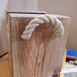 May include: A handcrafted wooden box with a natural wood grain finish. It features a thick, twisted rope handle. The box is rectangular and appears to be sturdy, suitable for storage or decorative purposes.