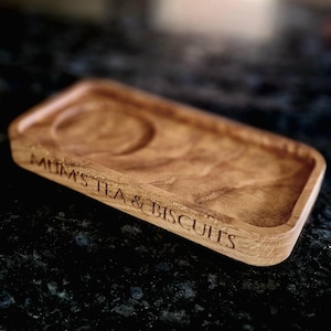 May include: A wooden tray with two indentations, one larger than the other. The tray is engraved with the words "Mum's Tea & Biscuits".