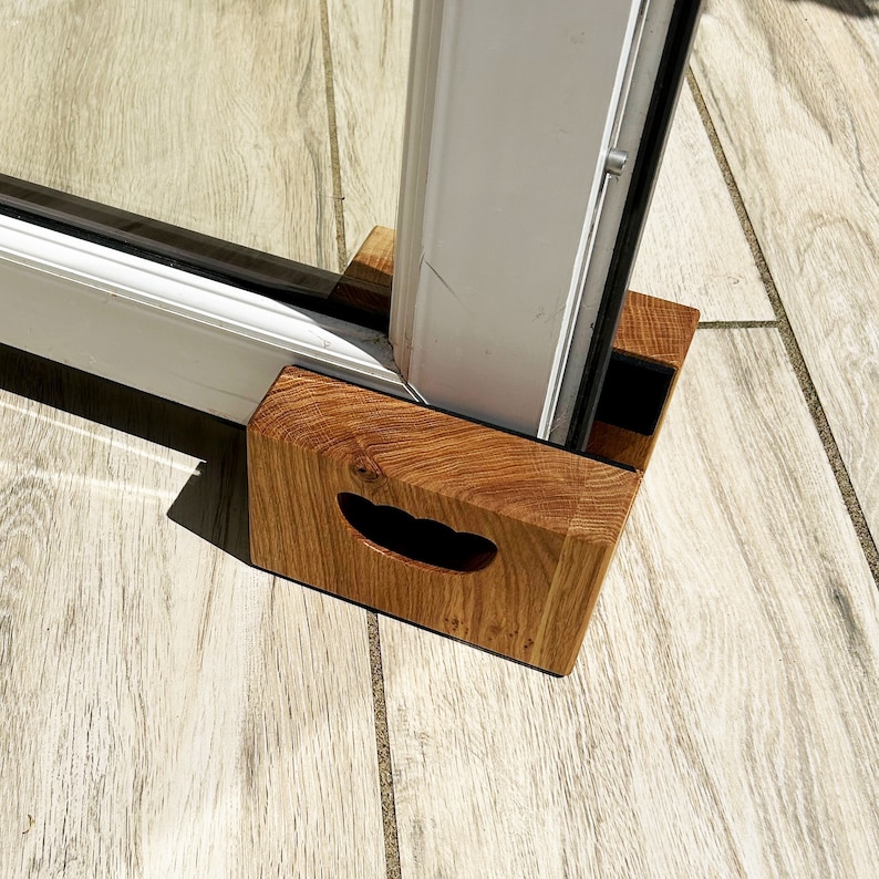 May include: A wooden door stop with a handle, positioned at the base of a white door frame. The door stop is made of light brown wood and has a rectangular shape with a curved handle cutout.