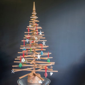 May include: A wooden Christmas tree decorated with colorful ornaments, including stars, hearts, and leaves. The tree is standing on a clear glass base.