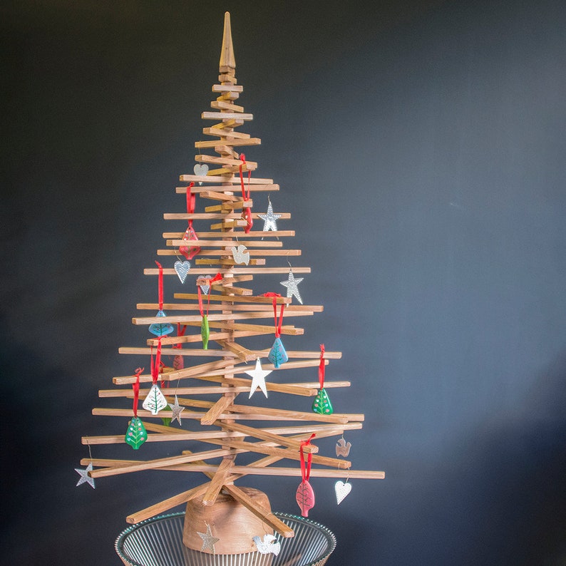 May include: A wooden Christmas tree decorated with colorful ornaments, including stars, hearts, and leaves. The tree is standing on a clear glass base.
