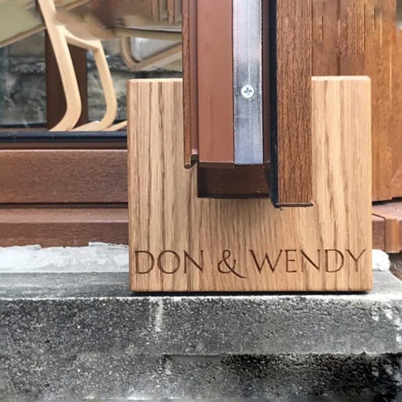 May include: A wooden plaque with the engraved text "DON & WENDY" is displayed. The plaque is made of light-colored wood and is positioned in front of a window frame. The window frame is brown and has a metal strip.