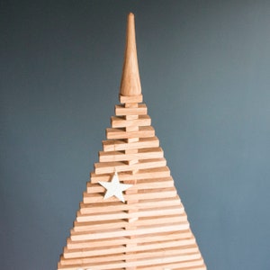 May include: A wooden Christmas tree made of stacked wooden slats with a star ornament hanging from the top. The tree is a light brown color and has a natural wood finish.