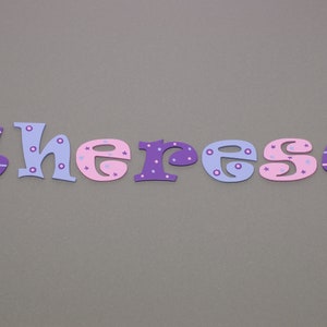 May include: The name "Theresa" spelled out in colorful, decorated wooden letters. The letters are purple, pink, and blue with white polka dots and stripes.