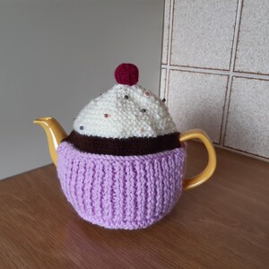 May include: A purple and white knitted teapot cosy shaped like a cupcake with a red cherry on top. The cosy is decorated with white and brown beads.