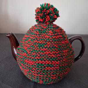 May include: A brown ceramic teapot with a hand-knitted tea cosy. The cosy is red and green, with a matching pom-pom. The teapot has a curved handle and spout.