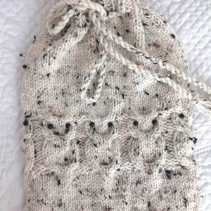 May include: A cream-colored knitted hot water bottle cover with a black speckled pattern and black beads around the bottom. The cover has a drawstring closure.