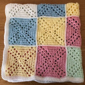 May include: A handmade crochet blanket featuring a patchwork design. The blanket is composed of square blocks in pastel colors: white, light blue, yellow, pink, and light green. A white border frames the blanket.