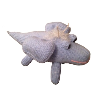 Puede incluir: Un animal de peluche de punto azul claro con forma de dragón con un cuerno blanco brillante y una boca rosa.