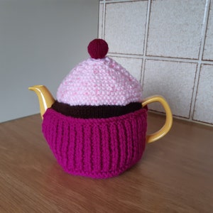 May include: A yellow teapot with a knitted pink and brown cupcake tea cosy. The cosy has a pink cherry on top.