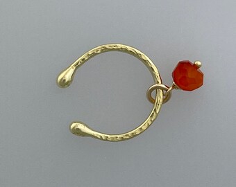 May include: A gold-colored ear cuff with a hammered texture. A small, faceted, orange-red gemstone hangs from the cuff. The ear cuff has a curved design with rounded ends, and is designed to be worn on the ear.