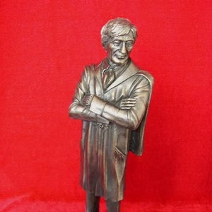 May include: A bronze sculpture of a man wearing a long coat and standing on a black base. The base has a gold plaque with the text "John Cleese,  Prof.  DeVere,  The  Hotel  Inspector,  1980".