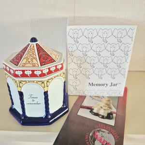 May include: A ceramic memory jar with a carousel design. The jar is white, red, and blue, with elephant motifs and the words "Times to remember." A box with elephant drawings and a brochure are also present.