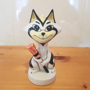 Puede incluir: Una figurita de cerámica de un gato blanco y negro sosteniendo una escoba roja y amarilla. El gato tiene una cara amarilla con ojos negros y una nariz negra. La figurita está sentada sobre una base blanca.