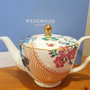 May include: A white teapot with a floral pattern and gold accents. The teapot has a lid with a gold knob and a spout on the side. The teapot is sitting on a wooden surface.