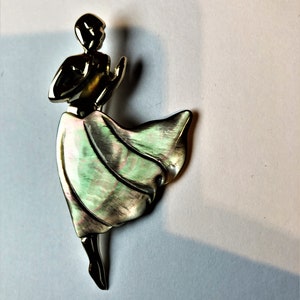 Pode incluir: Um broche dourado com uma mulher estilizada em um vestido verde e branco fluido. Os braços da mulher estão levantados e sua cabeça está inclinada para trás.