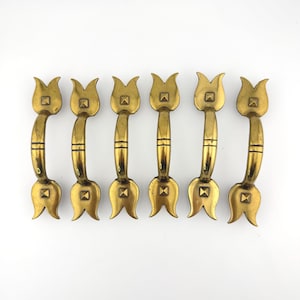 May include: A set of six brass drawer pulls, each featuring a curved handle with a central horizontal line detail and stylized tulip-shaped ends. Each end is accented with a small, raised square pyramid detail, creating a uniform metallic look.