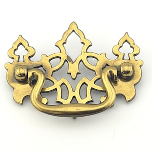 May include: A brass drawer pull featuring an ornate, openwork backplate with a symmetrical, cutout design and a curved, swinging handle attached to two circular mounting points. The metal has a warm, metallic gold finish with visible patina.