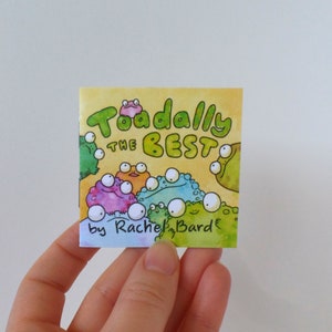 May include: A yellow book cover with the title "Toadally the Best" in green and black lettering. The cover features a group of cartoon frogs with large eyes and colorful bodies. The book is by Rachel Bard.