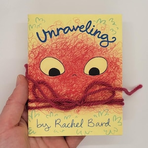 Op de afbeelding: Een geel boek met een rood en oranje pluche beest op de omslag. Het boek heet "Unraveling" en de auteur is "Rachel Bard".