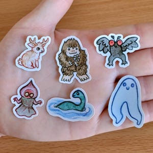 Puede incluir: Una colección de pegatinas caprichosas con varios críptidos y criaturas míticas. Los diseños incluyen un ciervo, Bigfoot sosteniendo una flor, un hombre polilla, un fantasma, una criatura de la laguna negra y el monstruo del lago Ness. Las pegatinas son coloridas y dibujadas a mano.