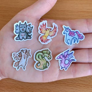 May include: A collection of seven fantasy creature stickers, including a three-headed dog, phoenix, unicorn, pegasus, hydra, and dragon. Each sticker features a unique, colorful design with black outlines and white borders. The stickers are displayed on a hand.