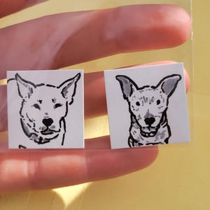 May include: Two small square stickers featuring black and white dog portraits. The dog illustrations have large ears and are in a minimalist style. The stickers are held in a clear plastic sleeve.