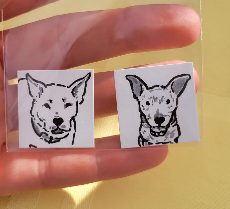 May include: Two small square stickers featuring black and white dog portraits. The dog illustrations have large ears and are in a minimalist style. The stickers are held in a clear plastic sleeve.